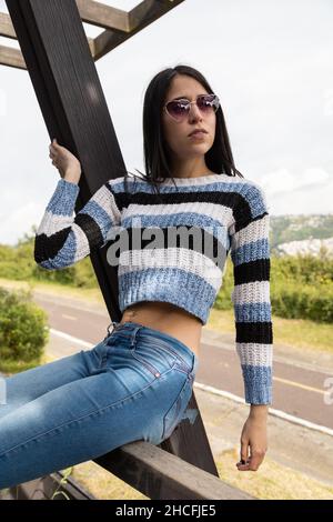 beautiful young latin female model sitting with black straight hair, wears sunglasses, jeans and casual sweater, beauty and fashion, lifestyle Stock Photo