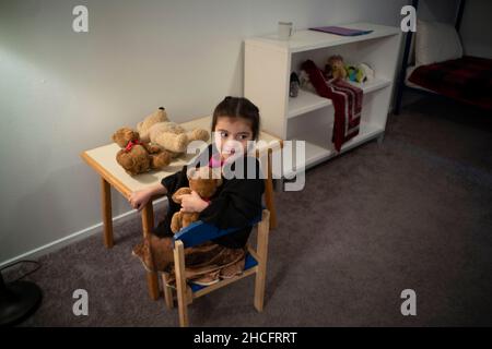 Austin Texas USA, December 2021: An Afghan child cuddles a stuffed bear and sits in a child-sized chair in the bedroom of her family's new apartment near downtown. Refugees fleeing turmoil in Afghanistan continue to be resettled in Texas with many single men and large families with children moving into apartments across Austin. Faith-based and other non-profit groups have stepped up to help coordinate efforts in furnishing homes for hundreds of refugees. ©Bob Daemmrich Stock Photo