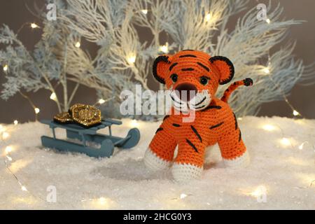 A knitted woolen tiger cub sits next to toy light blue trees with lights and toy sleds with coins on artificial white snow on a gray background. Close Stock Photo