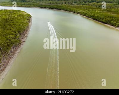 A power boat roaring up the river at speed leaving a wake behind disturbing the water. Tidal country river bordered by bushland. Stock Photo