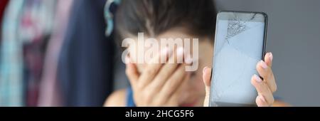 Sad woman covering her eyes with hand and holding broken mobile phone closeup Stock Photo