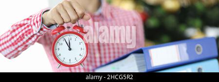 Woman holds alarm clock for twelve o'clock and stack of documents against background of Christmas tree Stock Photo
