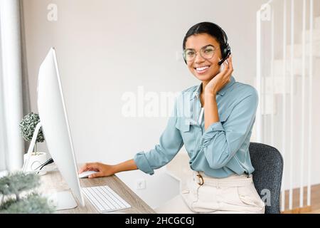 Young African-American woman wearing headset using computer for remote work while sitting at the home office. Sales woman making calls to customers, contacts online with clients Stock Photo