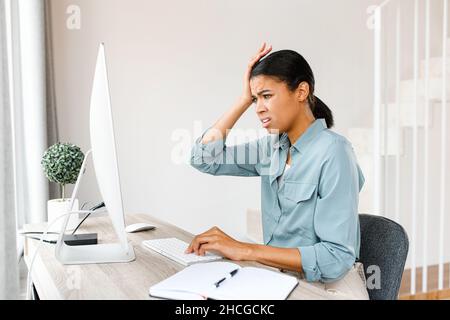 Tired African-American business woman sitting with computer at home and has a problem with remote work. Frustrated woman touching with hand to her head Stock Photo