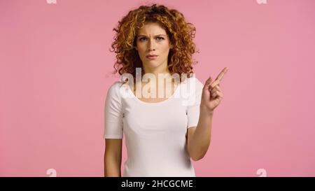 angry redhead woman looking at camera and pointing with finger isolated on pink Stock Photo