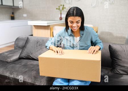Stylish multiracial brunette woman sitting on the couch with carton box on laps, received gift, feeling curious about ordered wished item from the website, satisfied with fast courier delivery service Stock Photo