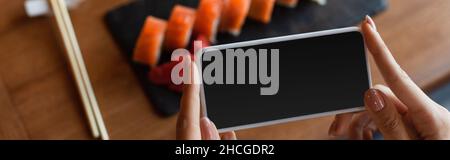 cropped view of woman holding smartphone with blank screen near blurred sushi rolls, banner Stock Photo