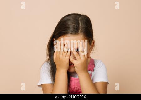 Little girl covering face with hands with slightly visible shining eyes and happy smile looking at camera wearing bright pink jumpsuit and white t Stock Photo