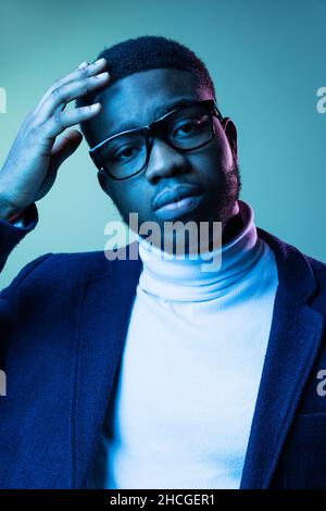 Close-up portrait of young stylish african man, student wearing white neck-polo and jacket isolated on blue background in neon light. Stock Photo