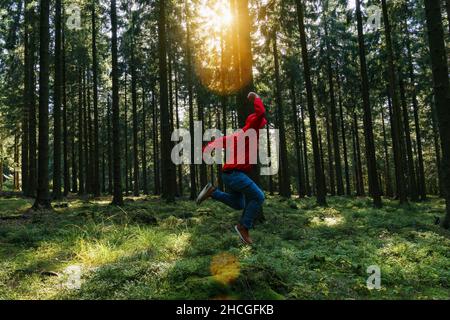 Young man jumps in the green forrest with red jacket and enjoys nature Stock Photo