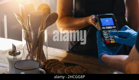 Waiter holding credit card swipe machine while customer with gloves typing Pin-code. Woman making payment in cafeteria with credit card. Customer pay Stock Photo
