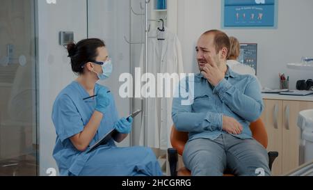 Dentistry nurse taking notes and talking to patient about toothache at oral care clinic. Woman assistant consulting man in pain with denture problems, discussing about dental procedure. Stock Photo
