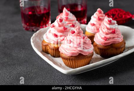 Homemade cupcakes decorated with cream and heart-shaped sprinkles for Valentine's Day in a heart-shaped plate on gray background Stock Photo