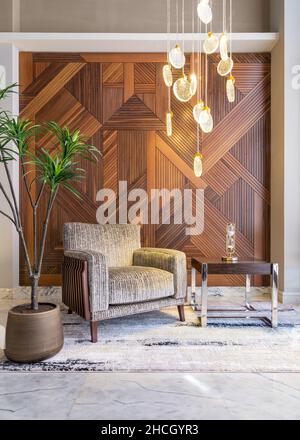 Modern beige armchair, small wooden modern table, planter with green bushes, and contemporary tall glass chandelier, in a hall with decorated wood cladding wall, and modern carpet on white marble wall Stock Photo