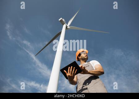 Indian engineer in orange helmet and grey overalls standing among windmill farm with modern tablet in hands. Foreman examining and controlling work of huge turbines. Stock Photo