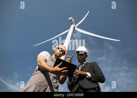 Indian engineer in uniform and helmet using tablet during meeting with african american inspector in stylish suit. Two partners discussing green energy while standing on farm with windmills. Stock Photo