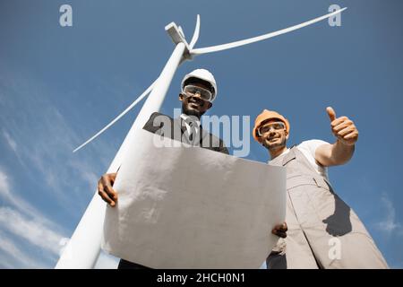 African american inspector with blueprints in hands having conversation with conception indian engineer among farm with wind turbines. Two colleagues discussing project outdoors. Stock Photo