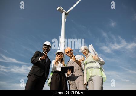 Group of four multiracial coworkers in safety helmets smiling on camera while staning on farm with huge windmills and showing thumbs up sign. Men and women using digital tablet and blueprints . Stock Photo