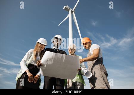Four multi ethnic partners in safety helmets studying wind turbine blueprints while standing together at rural area. Competent industrial workers discussing common project outdoors. Stock Photo