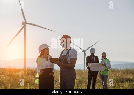 Team of multiracial industrial workers in safety helmets controlling process of green energy production on farm with wind turbines. Four colleagues using blueprints and tablet outdoors. Stock Photo