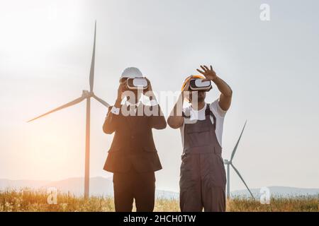 African american businessman and indian engineer wearing VR headset during meeting on farm with wind turbines. Innovative technologies and alternative energy concept. Stock Photo
