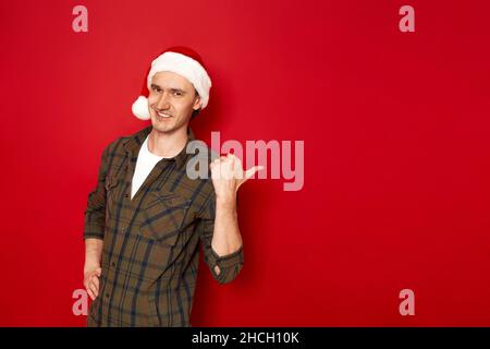 smiling man in Santa Claus Christmas hat, casual clothes points with his thumb at free empty area Stock Photo