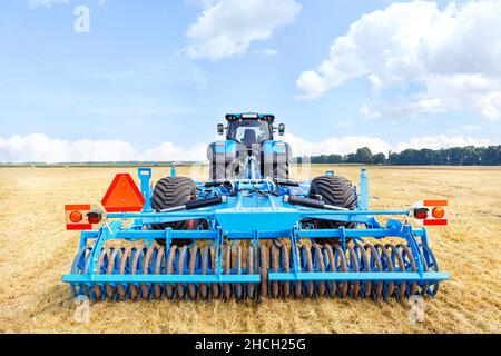 A trailed blue harrow on an agricultural tractor against the backdrop of a harvested yellow wheat field on a warm fall day. Central composition. Stock Photo