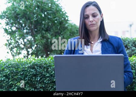 Beautiful Caucasian businesswoman using a laptop computer with a cup of coffee sitting outside an office building. Stock Photo