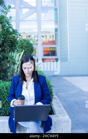 Beautiful Caucasian businesswoman using a laptop computer and holding a cup of coffee sitting outside an office building. Stock Photo