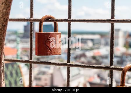 a rusted metal padlock hanging on a protective fence with a city view in a background Stock Photo