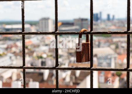 a rusted metal padlock hanging on a protective fence with a city view in a background Stock Photo