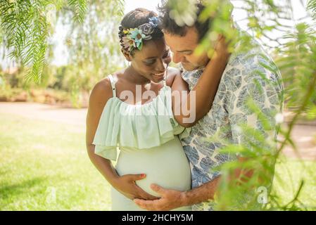 Happy diverse pregnant couple hugging and caressing belly in park Stock Photo