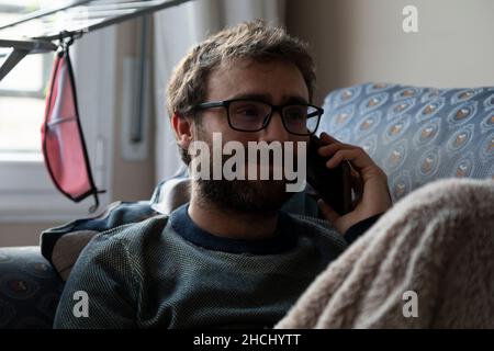 A young man, with pasta glasses, bearded, talks on the mobile phone with someone, while resting on the sofa, covered with a blanket, in the shared apa Stock Photo