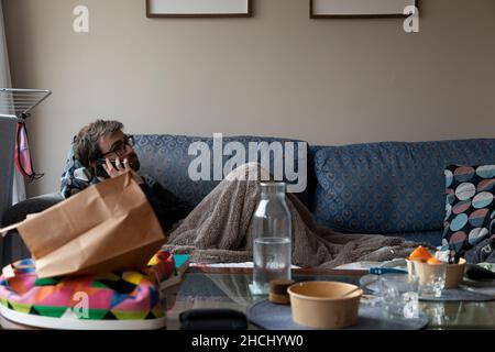 A young man, with pasta glasses, bearded, talks on the mobile phone with someone, while resting on the sofa, covered with a blanket, in the shared apa Stock Photo