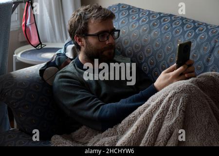 A young man with a beard surfs, chats and reads news on the internet with his smartphone, while he rests on the sofa, covered with a blanket, in the s Stock Photo
