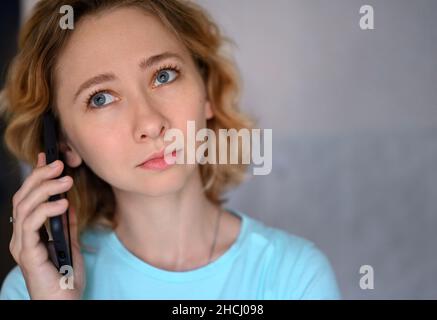 Close up portrait of young caucasian woman talking on cell phone at home.The blue-eyed blonde looks up thoughtfully. She is wearing a blue T-shirt. Stock Photo