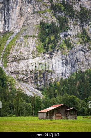 Small wooden house in a green grass field at the bottom of a massive rock wall in Lauterbrunnen valley, Switzerland, Europe Stock Photo