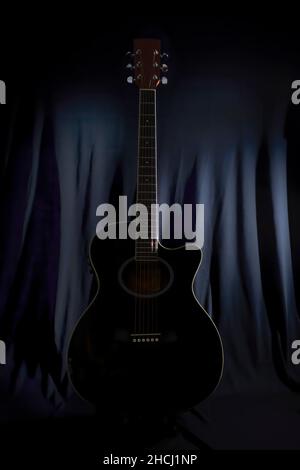 low key image of the silhouette of an acoustic guitar on a stage with a lightly illuminated black backdrop curtain in the background Stock Photo