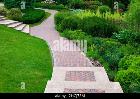 stone steps descent to a path from paving slabs in a park on a slope with ornamental plants, evergreen bushes and trees, a backyard landscape a place Stock Photo