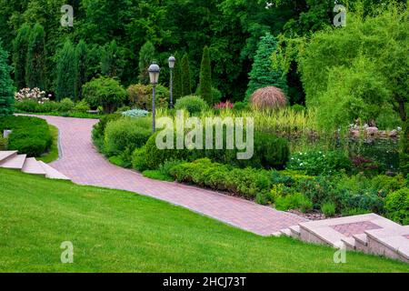 stone steps descent to a path from paving slabs in park with iron retro lantern on slope with scenic plants, evergreen bushes and pine and foliage tre Stock Photo