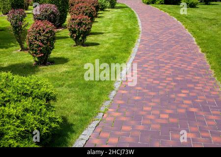curved stone tile pavement curved path in park landscaped with green grass and foliage bushes, garden landscape design of backyard on a sunny summer d Stock Photo