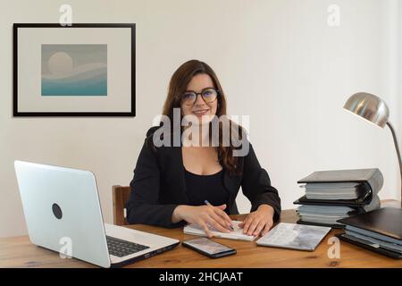 Beautiful Hispanic female executive office worker  smiling happily sitting at her desk with computer, smartphone and books looking forward against whi Stock Photo