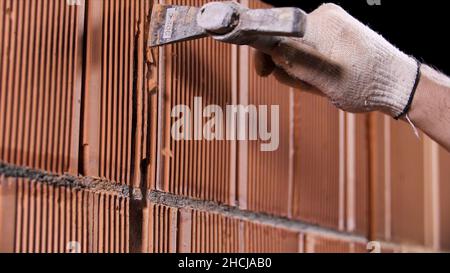 Close up of a man hammering the wall made of ceramic blocks at the construction site. Worker in protective gloves making a hole in the wall. Stock Photo