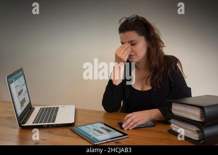 Beautiful Hispanic female office worker with headache massaging her eyes with her left hand, sitting at her desk in front of her computer Stock Photo