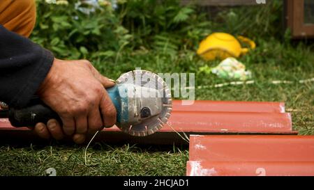 Roofer using an angle grinder machine to cut a roof tile of red color. Close up of male worker hands cutting a roof tile with a professional saw. Stock Photo