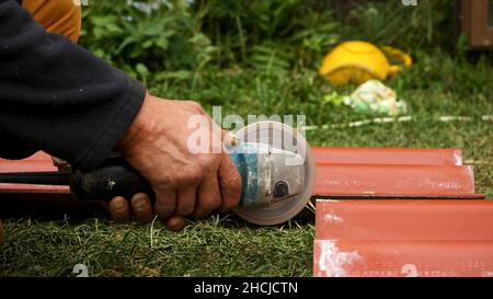 Roofer using an angle grinder machine to cut a roof tile of red color. Close up of male worker hands cutting a roof tile with a professional saw. Stock Photo
