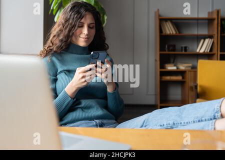 Young smiling woman holding smartphone chatting online while relaxing at cozy home office Stock Photo