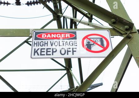 Photograph of a keep off high voltage safety sign on a large steel electrical transmission tower against a grey cloudy sky Stock Photo