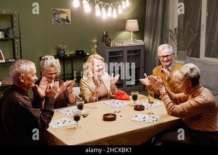 Three aged Caucasian women and two men sitting at wooden table with snack and drinks during party and playing board game Stock Photo