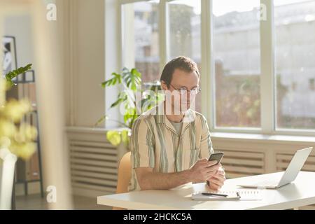 Horizontal medium shot of handsome Caucasian man wearing eyeglasses sitting at desk in modern office surfing Internet on smartphone Stock Photo